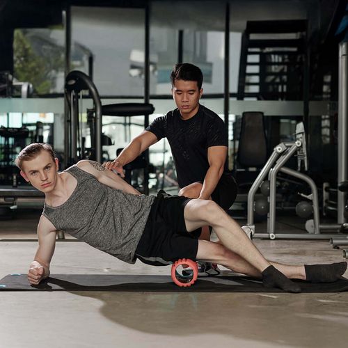 Junger Mann mit Trainer beim Training mit Faszienrolle im Fitnessstudio in Haslach