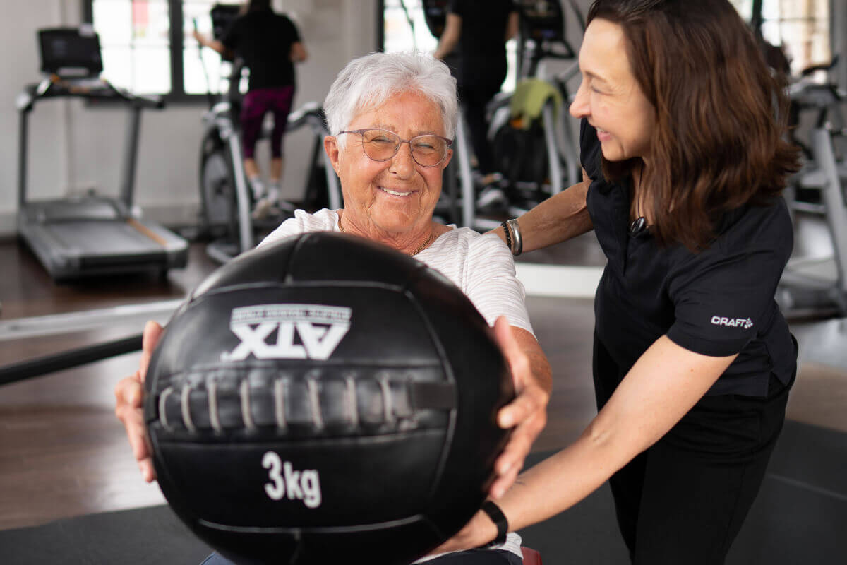 Ältere Dame mit Trainerin beim Training mit 3kg Ball im Fitnessstudio in Haslach